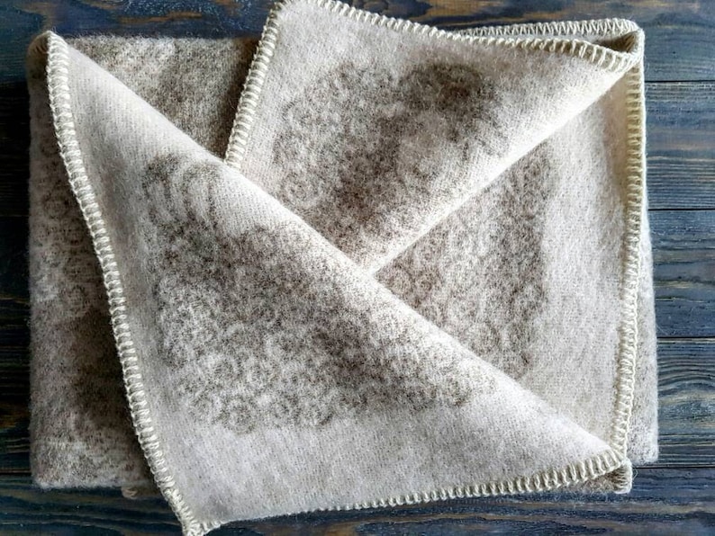 May include: A beige wool blanket with a brown floral pattern. The blanket is folded in half and has a white stitched border.