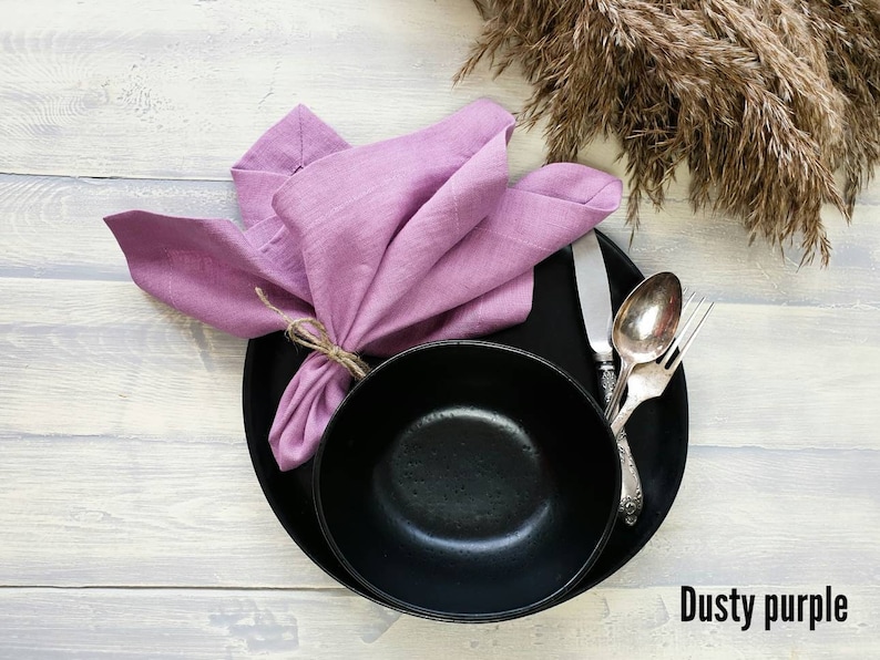 May include: A black bowl and plate with a dusty purple linen napkin on top. The napkin is tied with twine. A silver fork and spoon are on the plate.