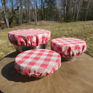 Puede incluir: Tres cubiertas de cuenco de tela a cuadros rojos y blancos de diferentes tamaños sobre una mesa de madera. Las cubiertas están hechas de una tela suave y transpirable y son perfectas para mantener los alimentos frescos.
