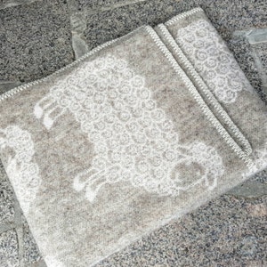 May include: A beige wool throw blanket with a white sheep design. The blanket has a fringe border and is folded in half.