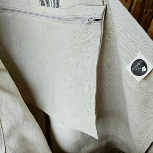 May include: A beige linen tote bag with a zipper closure and a striped accent. The bag has a long strap for carrying over the shoulder. The bag is made of a natural, textured fabric.