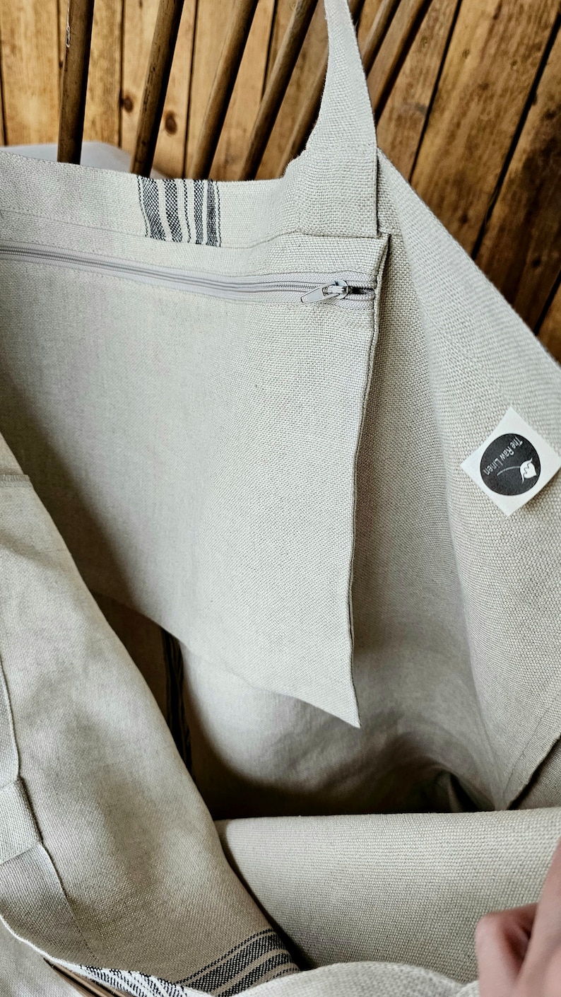 May include: A beige linen tote bag with a zipper closure and a striped accent. The bag has a long strap for carrying over the shoulder. The bag is made of a natural, textured fabric.