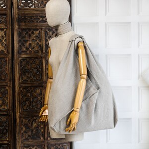 May include: A gray linen tote bag with a knotted handle, displayed on a wooden mannequin against a white paneled wall.