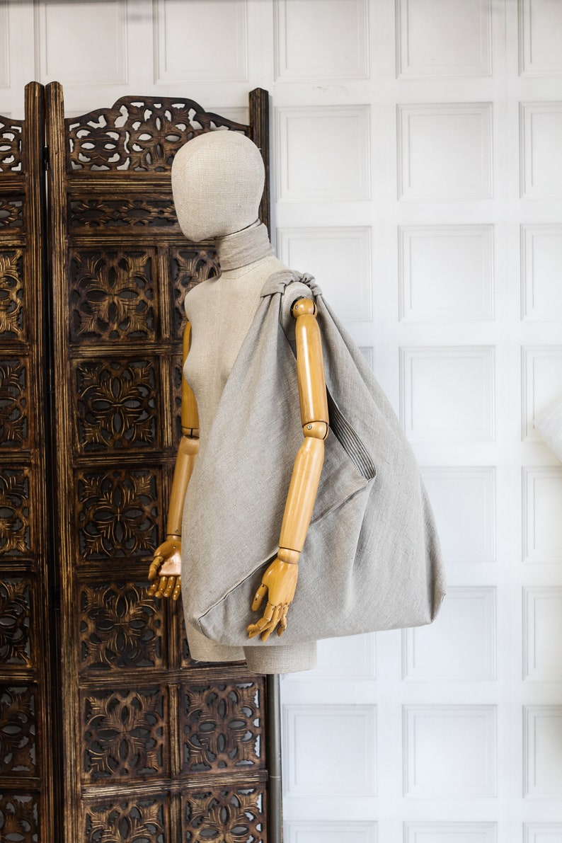 May include: A gray linen tote bag with a knotted handle, displayed on a wooden mannequin against a white paneled wall.