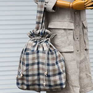May include: A large, brown and blue plaid tote bag with a drawstring closure and wooden bead accents. The bag is made of linen and has a casual, relaxed style.
