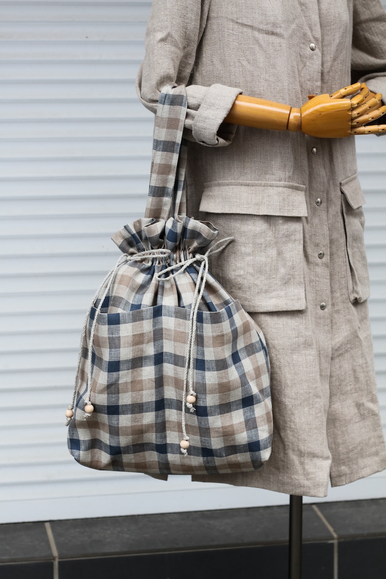 May include: A large, brown and blue plaid tote bag with a drawstring closure and wooden bead accents. The bag is made of linen and has a casual, relaxed style.