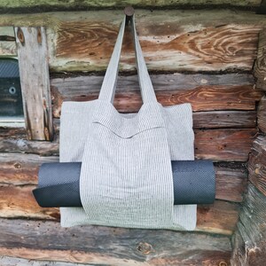 May include: A striped canvas tote bag with a black yoga mat tucked inside. The bag is hanging from a nail on a wooden wall.