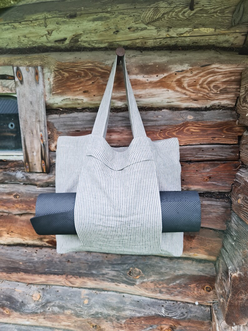 May include: A striped canvas tote bag with a black yoga mat tucked inside. The bag is hanging from a nail on a wooden wall.