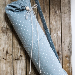 May include: A blue and white polka dot yoga mat bag with a drawstring closure and a long strap. The bag is hanging on a wooden wall.