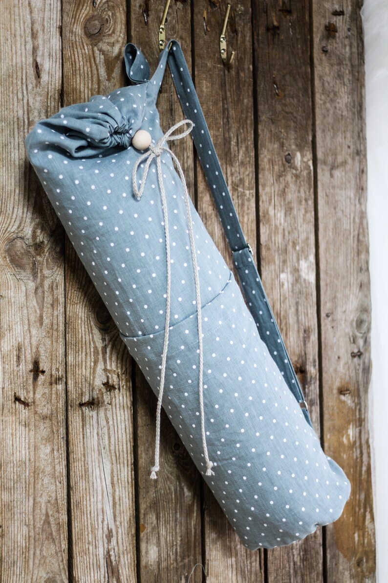 May include: A blue and white polka dot yoga mat bag with a drawstring closure and a long strap. The bag is hanging on a wooden wall.
