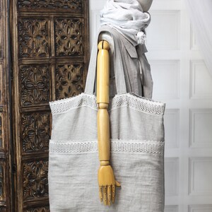 May include: A large, gray linen tote bag with white lace trim. The bag is being held by a wooden mannequin.