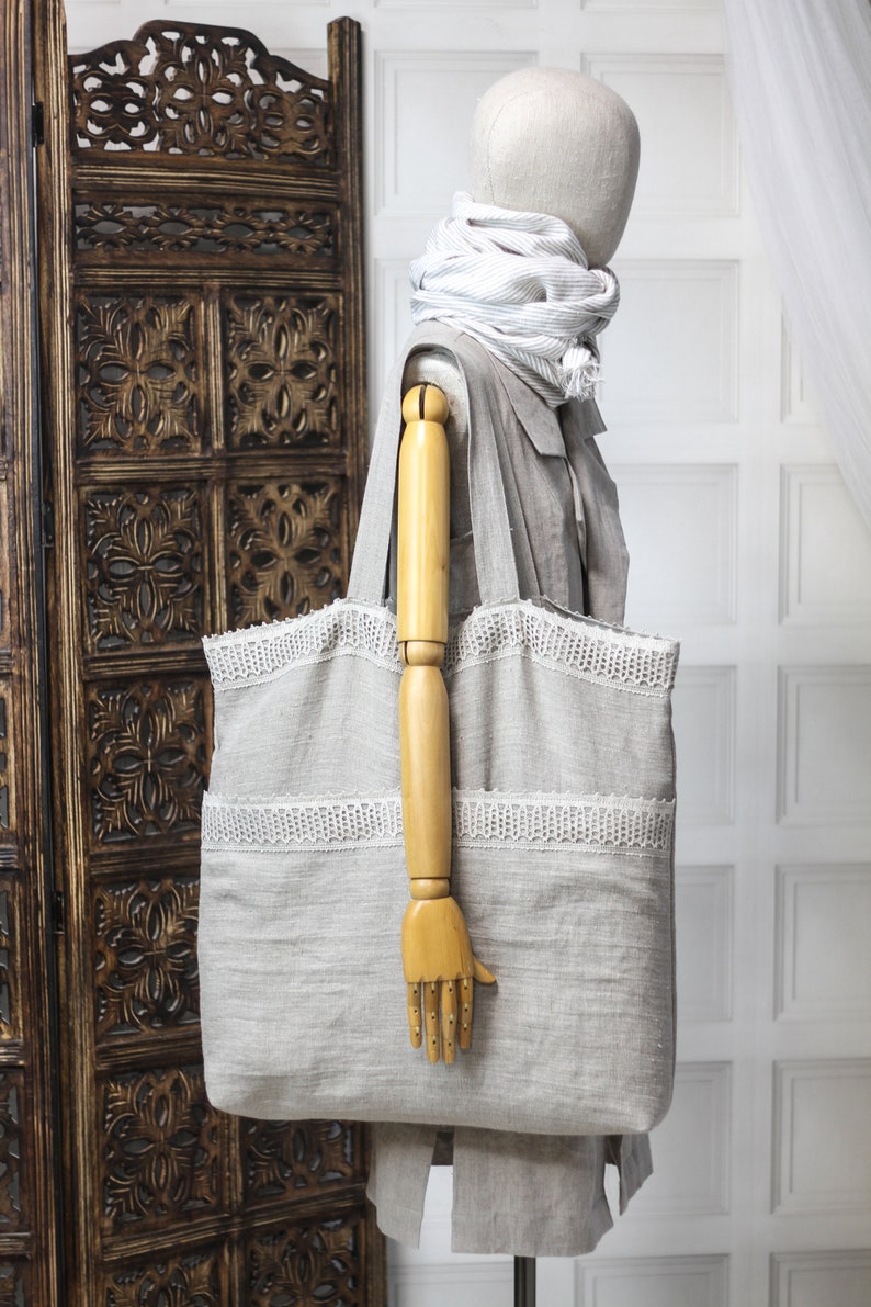 May include: A large, gray linen tote bag with white lace trim. The bag is being held by a wooden mannequin.