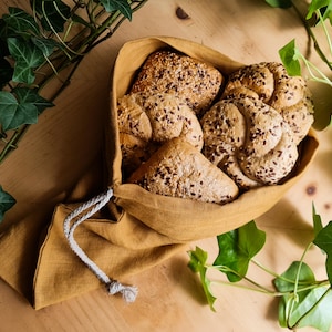 May include: A close-up of a brown linen bread bag filled with six round, seed-covered rolls. The bag is tied with a white string and sits on a wooden surface.