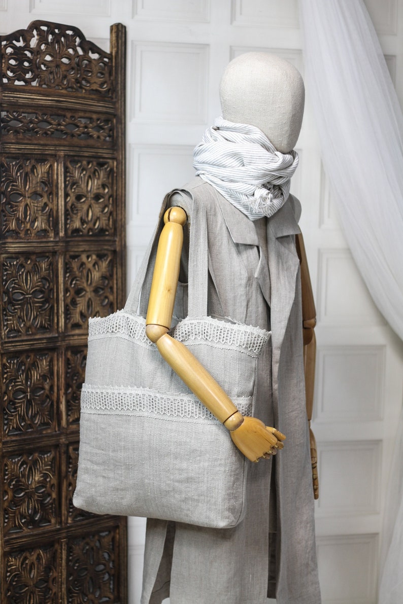 May include: A gray linen tote bag with white lace trim. The bag is being held by a wooden mannequin wearing a gray linen jacket and a white and gray striped scarf.