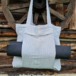 May include: A blue and white striped tote bag with a black yoga mat tucked inside. The bag has a pocket on the front and is hanging from a wooden wheel.