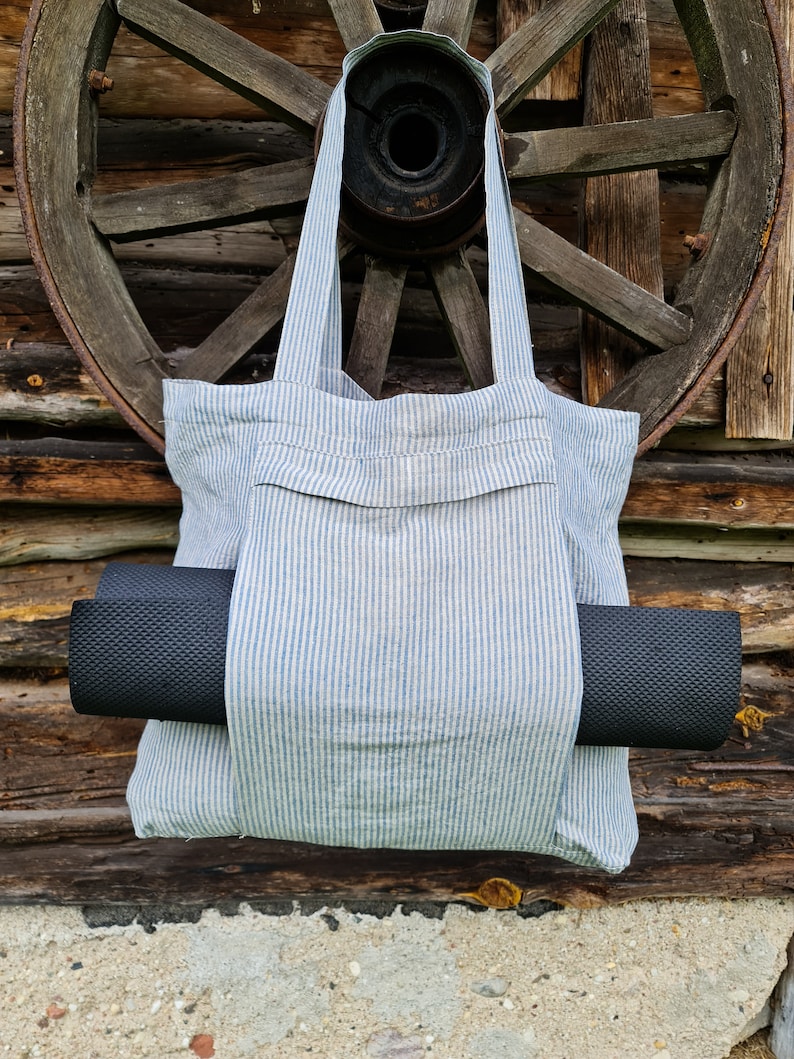 May include: A blue and white striped tote bag with a black yoga mat tucked inside. The bag has a pocket on the front and is hanging from a wooden wheel.
