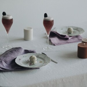 May include: A white tablecloth with two glasses of pink bubbly topped with blackberries, two candles, a plate with a white chocolate truffle, and two purple linen napkins.