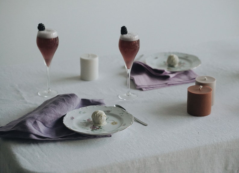 May include: A white tablecloth with two glasses of pink bubbly topped with blackberries, two candles, a plate with a white chocolate truffle, and two purple linen napkins.