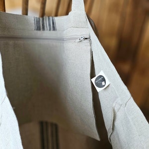 May include: Beige linen tote bag with a zipper closure and a striped design. The bag has long straps and a small tag with text. The bag is made of a natural-colored fabric.