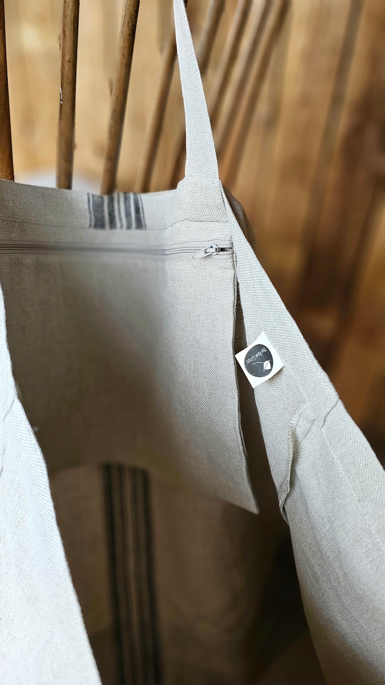 May include: Beige linen tote bag with a zipper closure and a striped design. The bag has long straps and a small tag with text. The bag is made of a natural-colored fabric.