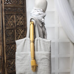 May include: A large, light gray tote bag with white lace trim. The bag is made of linen and has a wide, flat base. The bag is being held by a wooden mannequin.