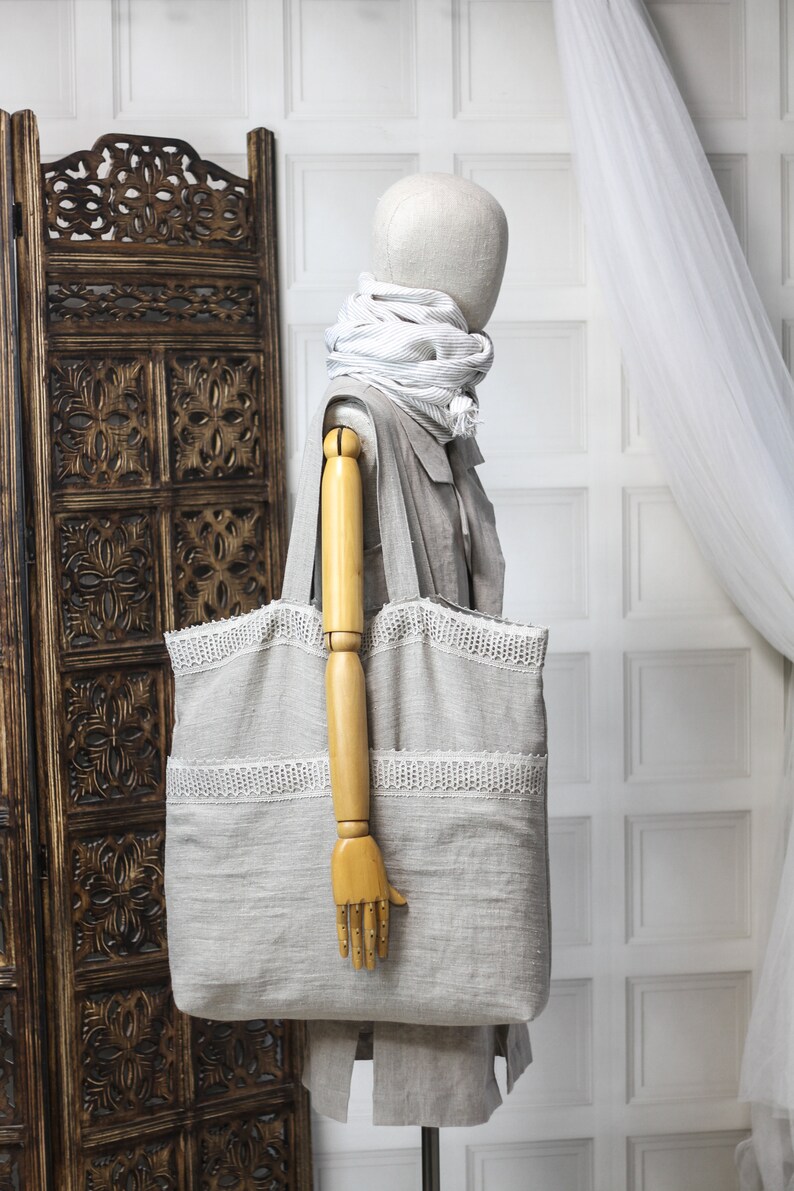 May include: A large, light gray tote bag with white lace trim. The bag is made of linen and has a wide, flat base. The bag is being held by a wooden mannequin.
