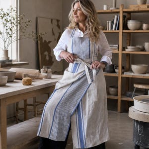 Puede incluir: Un delantal largo estilo envolvente hecho de lino beige natural con rayas verticales azules. El delantal tiene una cintura ancha que se ata al frente, una parte superior con peto y una falda larga con abertura lateral, sobre una camisa blanca.