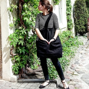 May include: A woman wearing a black and white striped shirt with a black apron over it. She is standing in front of a white wall with green vines growing on it.