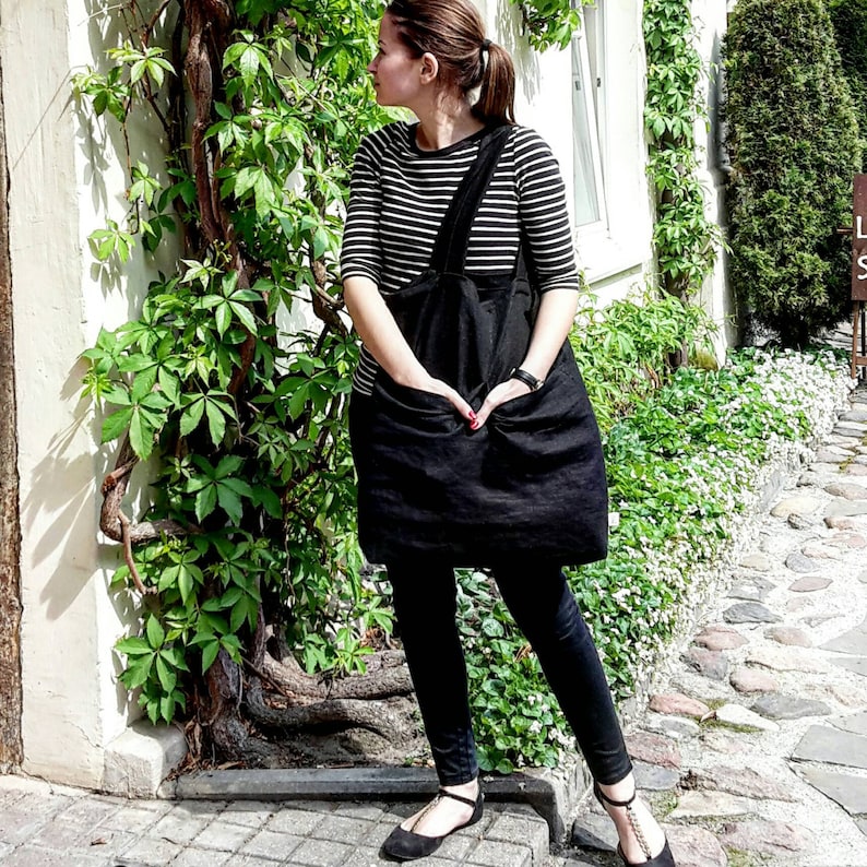 May include: A woman wearing a black and white striped shirt with a black apron over it. She is standing in front of a white wall with green vines growing on it.