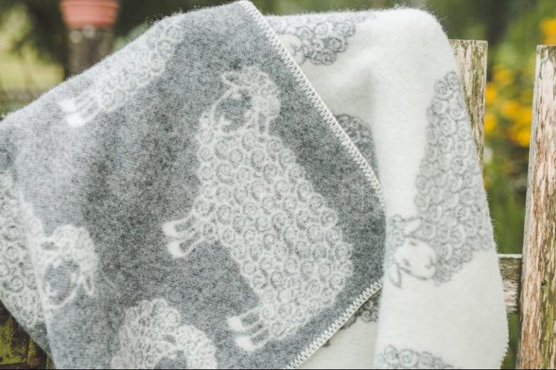 May include: A soft, white and gray throw blanket with a sheep pattern. The blanket is folded in half and draped over a wooden fence.