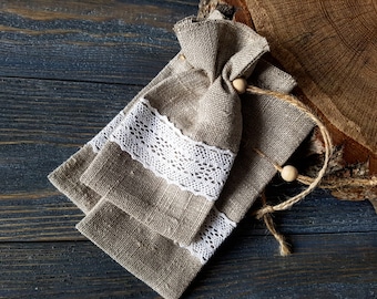 May include: Two small, beige linen bags with white lace trim and wooden beads. The bags are tied with twine and are lying on a dark wood surface.