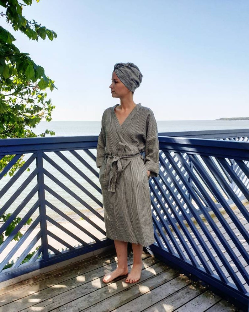 May include: A gray and white striped linen robe with a wrap closure and a matching head wrap. The robe is worn by a person standing on a wooden deck with a blue railing.