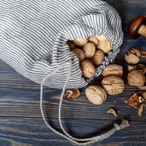 Può includere: Una borsa di lino a righe grigie e bianche piena di noci, con uno schiaccianoci di legno e noci aggiuntive sparse su una superficie di legno scuro.