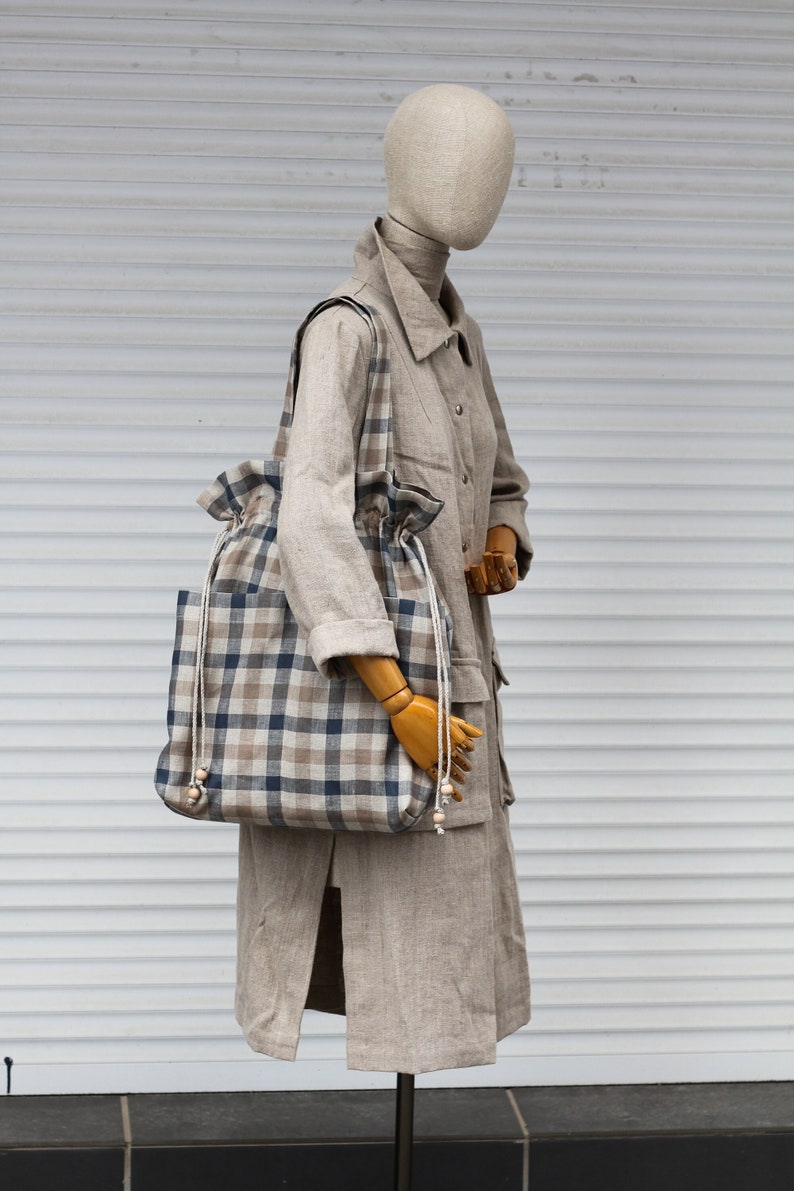 May include: A beige linen coat with a large plaid tote bag. The bag has a drawstring closure and is made of a natural fabric.