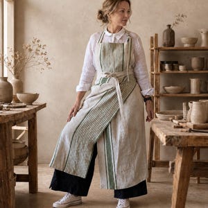 May include: A long, wrap-style apron made of natural-colored linen featuring vertical green stripes. The apron has a bib top, adjustable neck straps, and a waist tie that secures at the front, layered over a white long-sleeve shirt and dark pants.