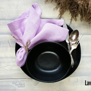 May include: A black bowl and plate with a lavender linen napkin tied with twine. The bowl and plate are on a light wood surface.