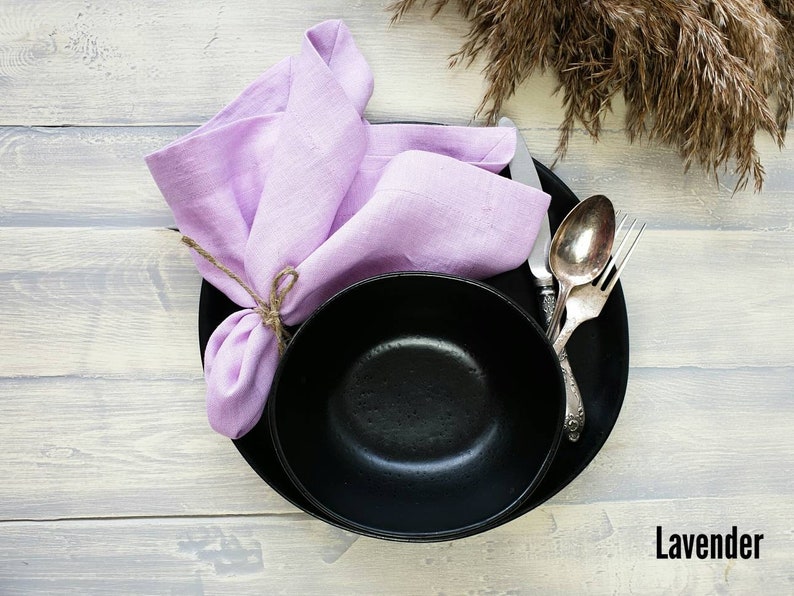 May include: A black bowl and plate with a lavender linen napkin tied with twine. The bowl and plate are on a light wood surface.