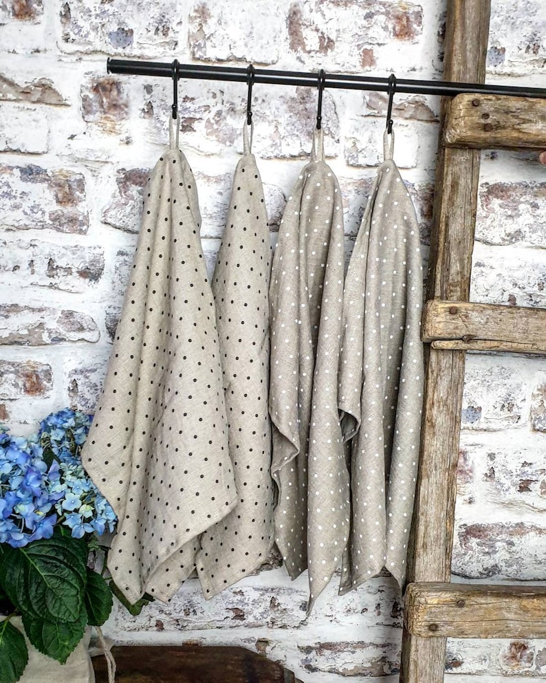 May include: Four beige linen tea towels with black polka dots hanging on a black metal rod. The towels are folded and hanging in a row.