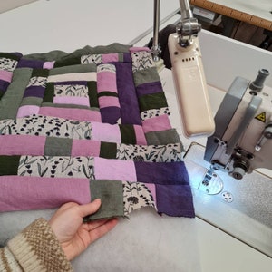 May include: A patchwork quilt top being sewn on a sewing machine. The quilt is made of various shades of pink, purple, and green fabric with a floral pattern. The quilt is being sewn with a white thread.