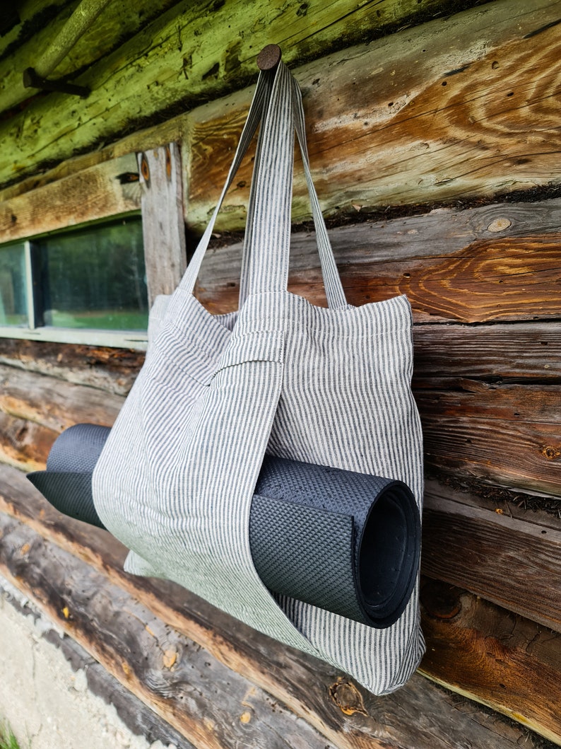 May include: A striped tote bag made of linen with a black yoga mat tucked inside. The bag is hanging on a wooden wall.