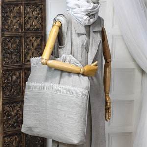 May include: A gray linen tote bag with a white lace trim. The bag is being held by a wooden mannequin wearing a gray linen jacket and a white and gray striped scarf.