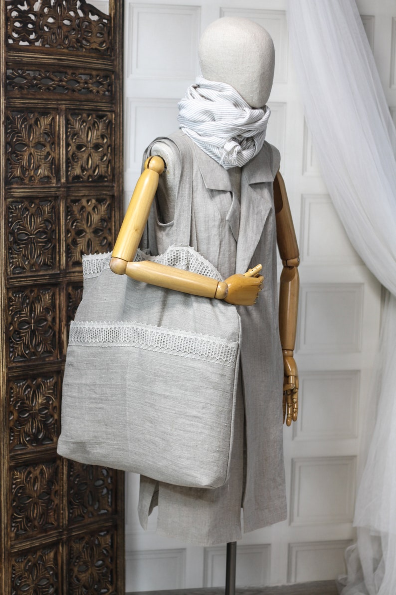 May include: A gray linen tote bag with a white lace trim. The bag is being held by a wooden mannequin wearing a gray linen jacket and a white and gray striped scarf.