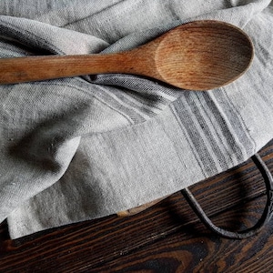 May include: A wooden spoon resting on a striped linen kitchen towel. The towel is folded and draped over a wooden surface.