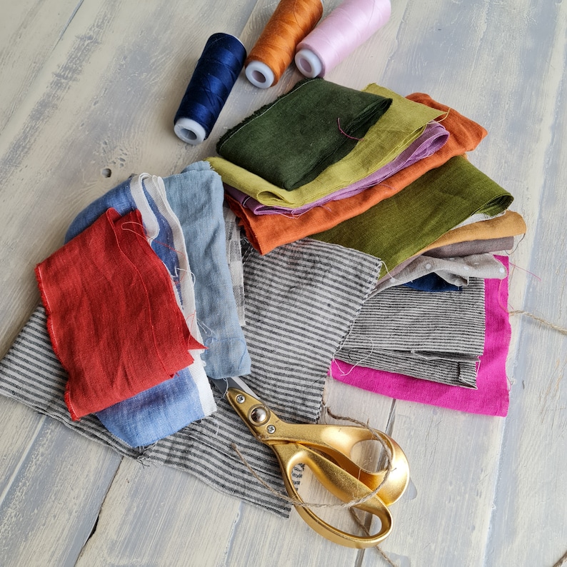 May include: A collection of fabric scraps in various colors and textures, including green, orange, blue, and red, with a pair of gold scissors. The fabric scraps are arranged in a pile, with some of the scraps folded over.