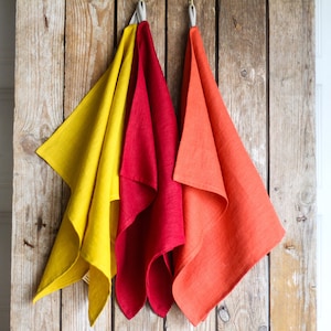 Puede incluir: Tres toallas de cocina de lino colgadas en una pared de madera. Las toallas son de color amarillo, rojo y naranja.