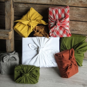 May include: A collection of seven fabric-wrapped gifts in various colors and patterns, including yellow, white, red, green, and brown. The gifts are arranged on a wooden surface.