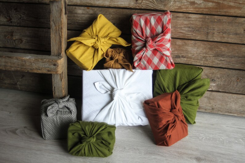 May include: A collection of seven fabric-wrapped gifts in various colors and patterns, including yellow, white, red, green, and brown. The gifts are arranged on a wooden surface.