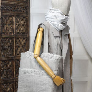 May include: A gray linen tote bag with white lace trim. The bag is being held by a wooden mannequin wearing a gray linen jacket and a striped scarf.