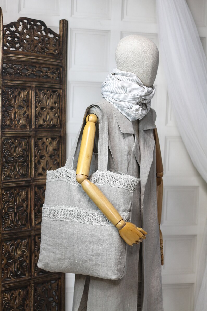 May include: A gray linen tote bag with white lace trim. The bag is being held by a wooden mannequin wearing a gray linen jacket and a striped scarf.