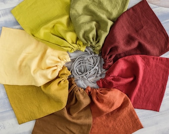 Puede incluir: Nueve bolsas de tela con cordón de ajuste en varios colores, incluyendo verde, amarillo, rojo, naranja y marrón. Las bolsas están dispuestas en círculo sobre una superficie de madera clara.